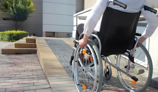 Eine Person in einem Rollstuhl fährt eine gepflasterte Rampe hinauf. Die Umgebung besteht aus modernen Gebäuden und grünen Pflanzen.