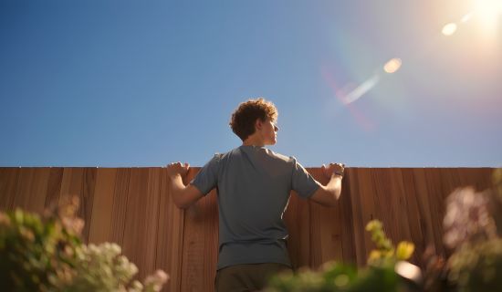 Ein Mann mit lockigen Haaren blickt über einen hohen Holzlattenzaun. Er steht im Gegenlicht der Sonne, der Himmel ist klar blau.