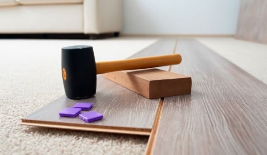 Das Bild zeigt eine Nahaufnahme von Werkzeugen und Material zur Laminatverlegung: Ein schwarzer Gummihammer, ein Schlagklotz und mehrere helle Laminatdielen liegen auf einem beigefarbenen Teppichboden. Im unscharfen Hintergrund ist ein häuslicher Wohnraum