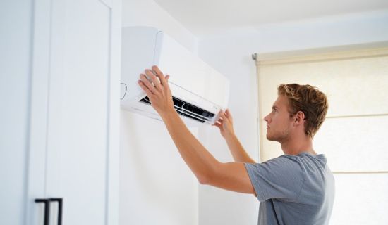 Ein junger Mann in einem grauen T-Shirt hängt vorsichtig das weiße Innengerät einer Klimaanlage in die dafür vorgesehene Wandhalterung. Er wird in einem hellen, modernen Raum von der Seite gezeigt.