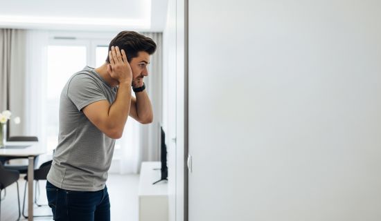 In einem hellen, modernen Raum steht ein Mann und hält sich mit beiden Händen die Ohren zu. Im Hintergrund sind Teile einer Wohnungseinrichtung wie ein Esstisch und ein Fenster zu erkennen. Seine Geste vermittelt eine Atmosphäre von Unbehagen oder dem Wun