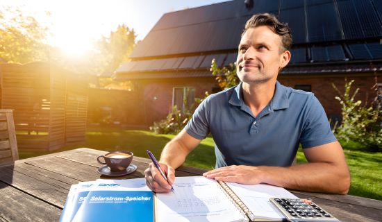 Ein Mann sitzt an einem sonnigen Gartentisch und macht sich mit Notizbuch und Taschenrechner an die Planung seiner Solaranlage. Im Hintergrund ist sein Haus mit Photovoltaik-Modulen auf dem Dach zu sehen.
