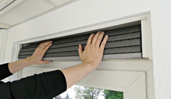 Zwei Hände schieben eine flexible, dunkelgraue und segmentierte Dämmmatte in die Öffnung eines weißen Rollladenkastens. Die Aktion zeigt das Einsetzen der Dämmung über einem Fenster als letzten Schritt der Isolierung.