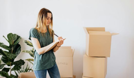 Frau notiert sich Dinge auf einem Block, hinter ihr stehen Umzugskartons