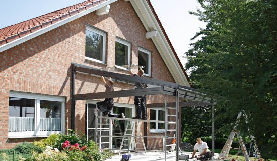 Handwerker montieren ein Glas-Terrassendach an einem roten Backsteinhaus. Zwei Arbeiter stehen auf einem Gerüst und befestigen Glasplatten an der Metallkonstruktion, während ein weiterer Arbeiter am Boden arbeitet. Im Vordergrund blühen rote Rosen in eine