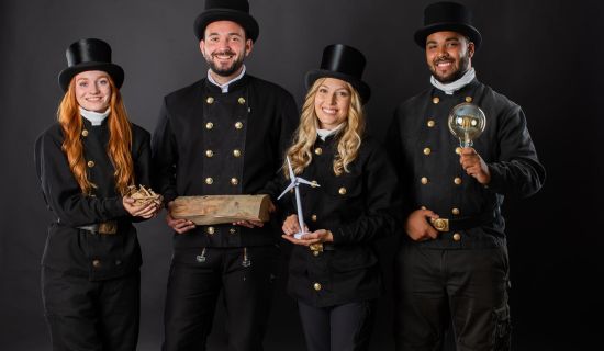 Vier Schornsteinfeger stehen vor einem schwarzen Hintergrund, gekleidet in schwarze Anzüge mit goldenen Knöpfen und Zylinderhüten. Sie halten verschiedene Objekte in ihren Händen, darunter ein Holzstück, eine Glühbirne und ein Windrad.