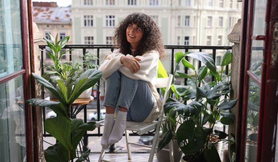 Frau sitzt auf einem Balkon mit vielen Pflanzen und schaut fröhlich-verträumt