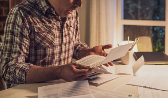 Eine Person in einem karierten Hemd sitzt an einem Tisch, umgeben von zahlreichen Dokumenten und Notizen, während sie konzentriert in mehreren Blättern blättert. Der Raum ist schwach beleuchtet und vermittelt eine ruhige Arbeitsatmosphäre.