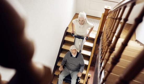 Ältere Frau fährt ihren Partner im Treppensteiger eine Holztreppe im Haus hinunter. Beide wirken entspannt.