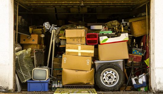 Ein überfülltes Garagentor zeigt eine chaotische Ansammlung von Kartons, Möbeln, einem alten Fernseher und verschiedenen Gegenständen, die den Raum nahezu vollständig ausfüllen. Die Szene vermittelt den Eindruck von Unordnung und Vernachlässigung.