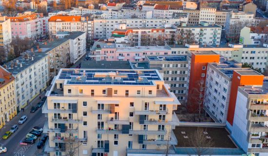 Luftaufnahme eines Stadtviertels mit modernen Wohngebäuden, einige mit Solarpaneelen auf den Dächern. Die Gebäude sind dicht bebaut, und die Sonne taucht die Szene in ein warmes Licht.