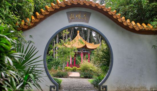 Ein traditionelles chinesisches Mondtor, eine runde Öffnung in einer weißen Gartenmauer, gibt den Blick frei auf einen Pagoden-Pavillon im Hintergrund. Üppige grüne Pflanzen und Bambus umrahmen die friedliche Szene.