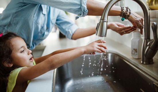 Ein kleines Mädchen mit dunklen Haaren und einem gelben Oberteil streckt seine Hände unter einen laufenden Wasserhahn in einem Küchenwaschbecken. Die Hände einer erwachsenen Person daneben betätigen einen Seifenspender. Das Bild stellt die alltägliche Nut