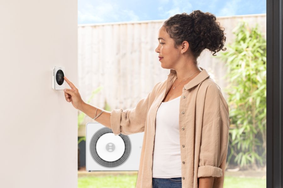 Eine Frau bedient ein rundes, digitales Raumthermostat an einer weißen Innenwand. Durch die offene Tür im Hintergrund ist im Garten eine moderne Wärmepumpen-Außeneinheit unter klarem Himmel zu sehen.