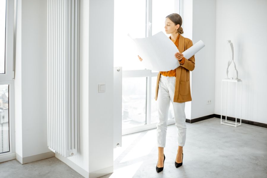 Eine Frau steht in einem leeren, lichtdurchfluteten Raum und betrachtet große Baupläne, die sie in den Händen hält. Der Raum verfügt über weiße Wände, einen Heizkörper und große Fensterfronten.