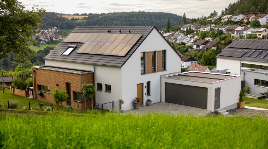 Modernes Einfamilienhaus mit weißen Wänden, Holzverkleidung und Solarpaneelen auf dem Dach, umgeben von grünem Gras und weiteren Häusern in hügeliger Landschaft unter bewölktem Himmel.