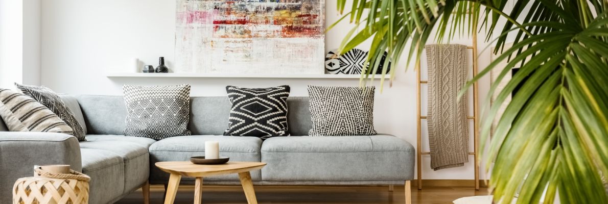 Ein helles, modernes Wohnzimmer mit einem großen grauen Ecksofa und verschiedenen schwarz-weiß gemusterten Kissen. Ein runder Jute-Pouf liegt auf einem hellen Teppich vor einem niedrigen Holztisch, während eine große Palme rechts den Raum mit Grün belebt.
