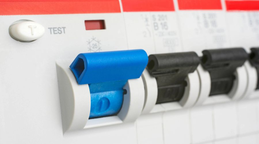 Nahaufnahme eines geöffneten Sicherungskastens mit Fokus auf einen blauen Fehlerstrom-Schutzschalter. Daneben befindet sich die weiße Test-Taste, während rechts weitere schwarze Leitungsschutzschalter aufgereiht sind.