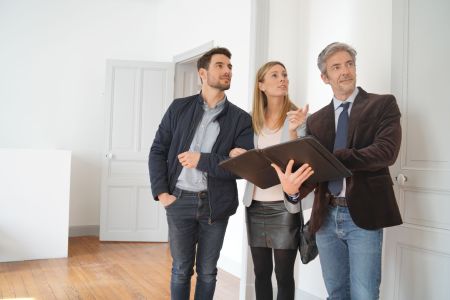 Ein Immobilienmakler im braunen Sakko hält eine Dokumentenmappe und deutet mit erhobener Hand in einen hellen, unmöblierten Raum mit Holzboden. Neben ihm steht ein interessiertes junges Paar, das seinen Ausführungen folgt. Im Hintergrund sind weiße Türen