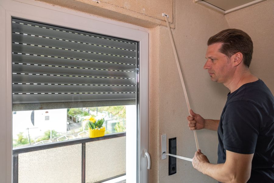 Ein Mann in einem schwarzen T-Shirt steht in einem Raum neben einem Fenster und betätigt den weißen Gurt des Rollladens. Der Gurtwickler und der Gurtschlitz in der Wand sind oft Quellen für unangenehme Zugluft.