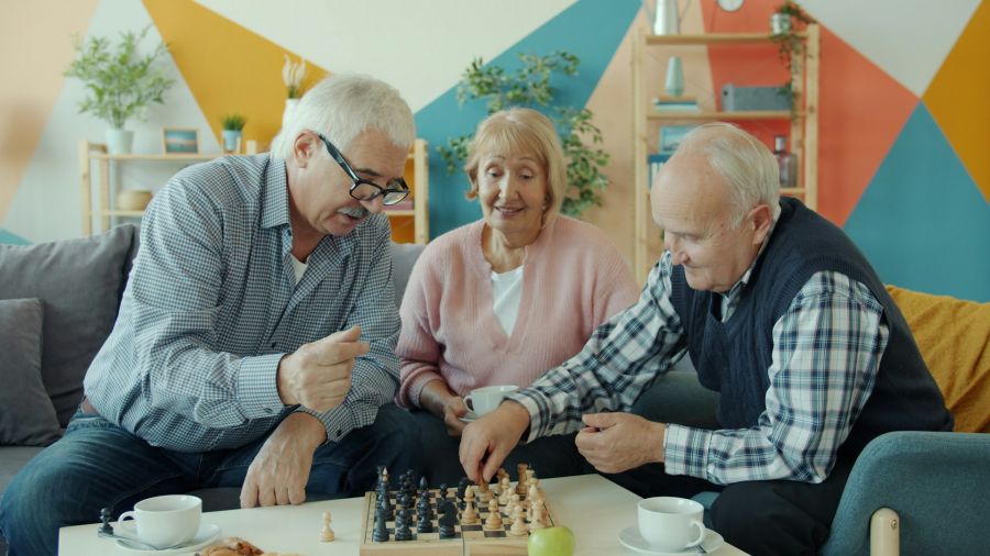Drei Senioren - zwei Männer und eine Frau - sitzen gemeinsam im Wohnzimmer und spielen Schach.