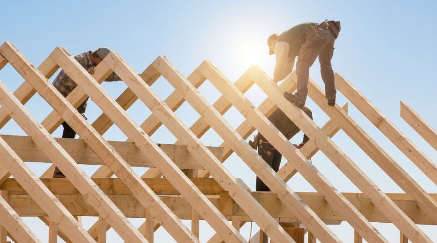 Handwerker in Arbeitskleidung balancieren auf den hölzernen Balken eines offenen Dachstuhls unter strahlend blauem Himmel und montieren die Konstruktion.