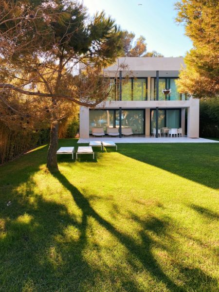Ein sehr modernes, zweistöckiges und kubisches Einfamilienhaus mit großflächigen Glasfronten strahlt im Sonnenlicht. Davor erstreckt sich eine makellos gepflegte, leuchtend grüne Rasenfläche. Im Vordergrund wirft ein großer Nadelbaum einen markanten Schat