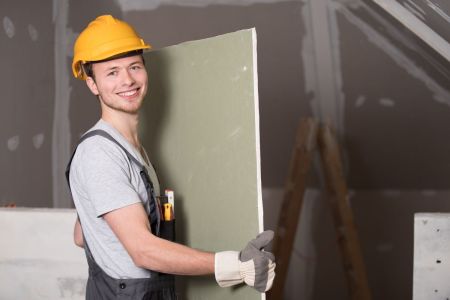 Hier ist ein junger Handwerker mit gelbem Schutzhelm und Arbeitskleidung auf einer Baustelle zu sehen, der eine Gipskartonplatte hält. Er lächelt zufrieden und zeigt den Daumen hoch, was eine positive und zuversichtliche Arbeitsatmosphäre vermittelt. Die