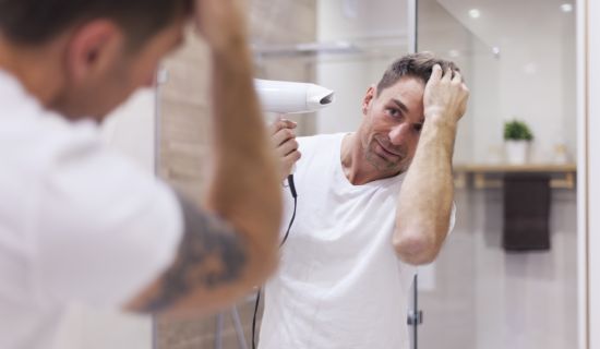 Ein Mann in einem weißen T-Shirt steht im Badezimmer vor dem Spiegel und föhnt sich die Haare. Er betrachtet sein Spiegelbild, das klar zu erkennen ist, was auf eine gute Belüftung hindeutet.