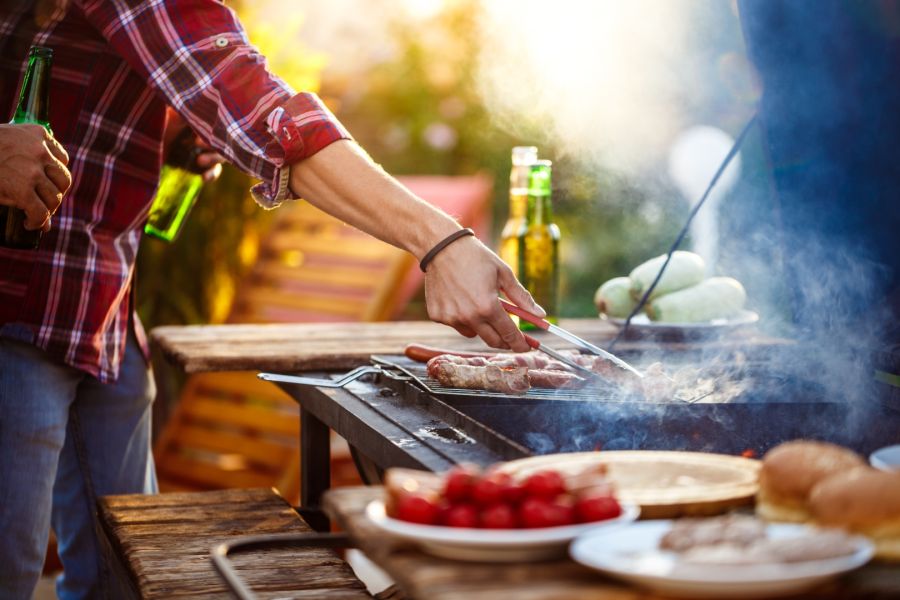 Nahaufnahme einer Person, die mit einer Grillzange Fleisch auf einem Holzkohlegrill im Freien wendet. Daneben steht eine weitere Person mit Bierflaschen in der Hand, während Rauch im Sonnenlicht aufsteigt.