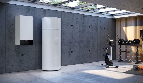 Eine weiße, wandhängende Wärmepumpen-Inneneinheit installiert neben einem hohen, zylindrischen Pufferspeicher in einem modern gestalteten Kellerraum. Der Raum verfügt über Sichtbetonwände, ein Oberlicht und ist teilweise als Fitnessraum mit Hantelbank und