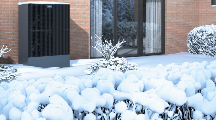 Eine moderne, dunkle Wärmepumpen-Außeneinheit steht neben einer Ziegelwand im verschneiten Garten vor einer großen Fensterfront. Die umliegenden Sträucher sind tief verschneit.