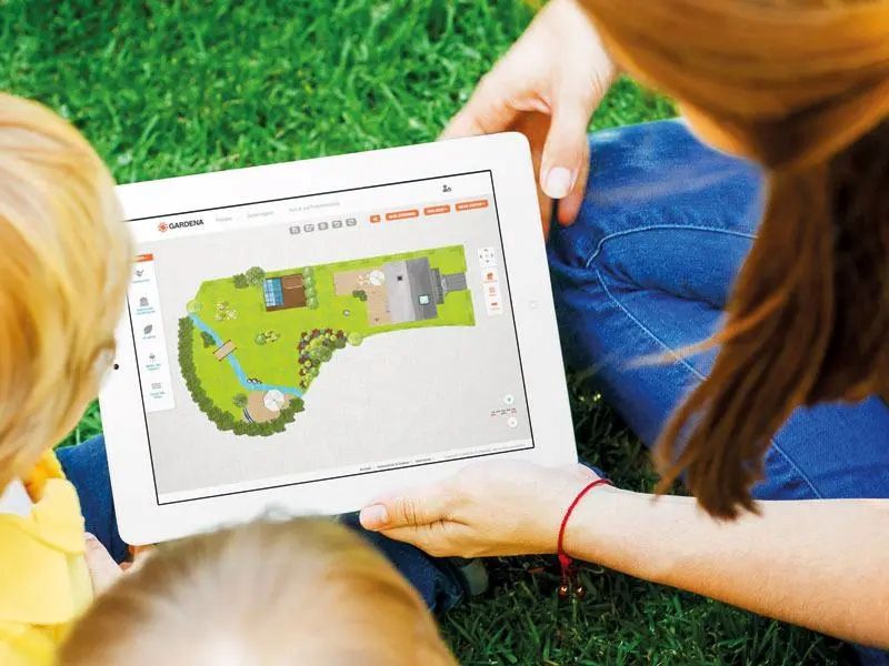 Eine Frau und zwei Kinder betrachten auf einem Tablet einen digitalen Gartenplan mit Rasen, Beeten und Wegen.