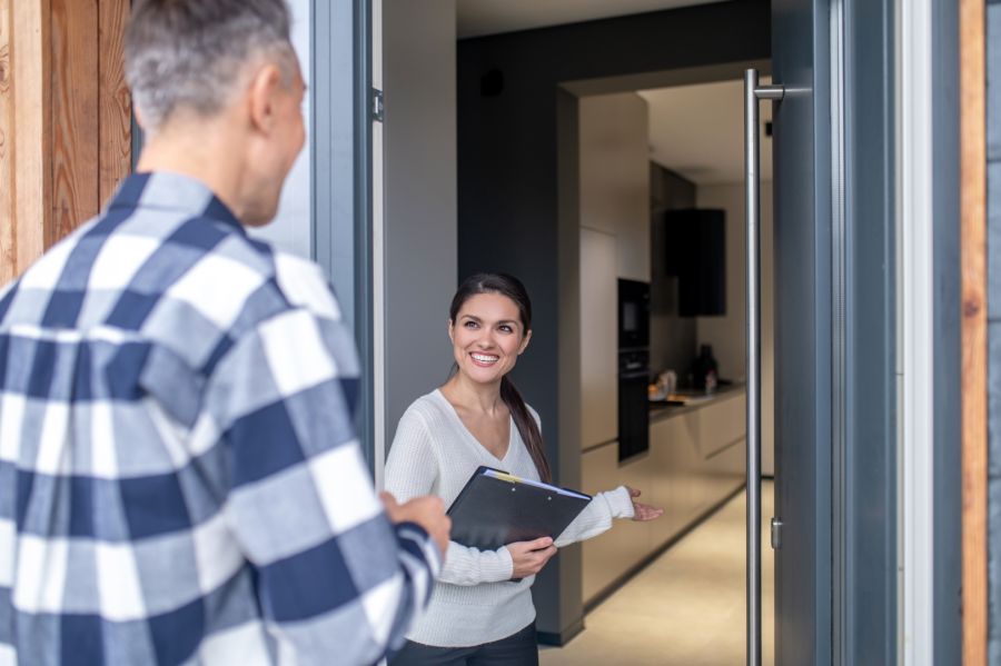 Eine freundlich lächelnde Immobilienmaklerin mit einem Klemmbrett in der Hand steht in einer geöffneten Wohnungstür und bittet einen Mann herein. Im Hintergrund sind der Flur und eine moderne Einbauküche zu sehen.