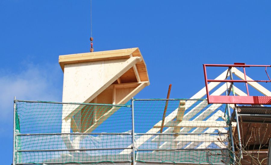 Eine große, vorgefertigte hölzerne Dachgaube hängt an einem Kranhaken über einer Baustelle mit grünem Sicherheitsnetz vor blauem Himmel.