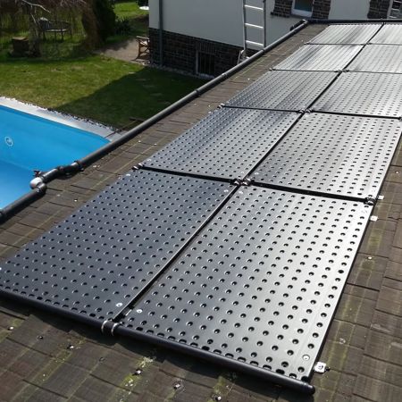 Schwarze Solarabsorber-Paneele sind auf einem dunklen Ziegeldach montiert, um das Poolwasser direkt mit Sonnenenergie zu erwärmen. Im Hintergrund ist ein Teil eines Pools mit einer blauen Abdeckung zu sehen, was die direkte Verbindung zwischen der Solaran