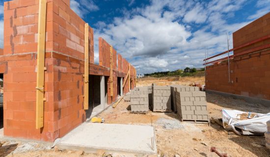 Eine Baustelle im Rohbauzustand unter blauem Himmel mit Wolken. Zu sehen sind halbfertige Wände aus roten Ziegelsteinen, Betonfundamente und Paletten mit grauen Baumaterialien auf dem sandigen Boden.