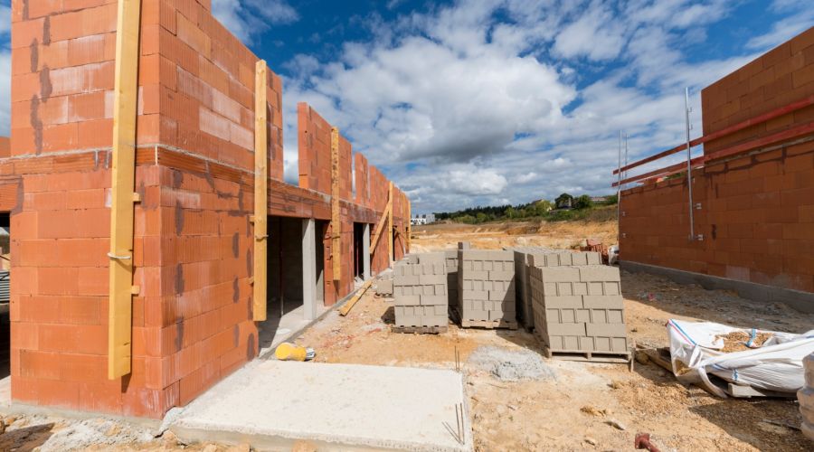 Eine Baustelle im Rohbauzustand unter blauem Himmel mit Wolken. Zu sehen sind halbfertige Wände aus roten Ziegelsteinen, Betonfundamente und Paletten mit grauen Baumaterialien auf dem sandigen Boden.