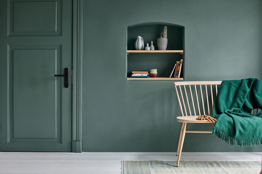 Ein modern eingerichteter Raum mit einer dunkel-salbeigrünen Wand und einer exakt farbgleichen Kassettentür. Rechts im Bild steht ein heller Holzstuhl mit einer grünen Decke, daneben eine Wandnische mit Büchern und Dekoration.