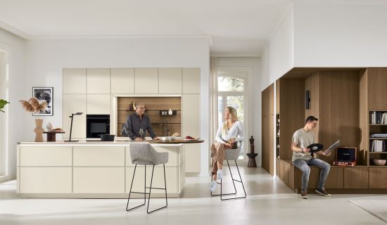 Eine helle, moderne Küche mit einer großen Kochinsel in Beige und Holzoptik. Drei Personen beleben den Raum: Ein Mann steht an der Insel, eine Frau sitzt auf einem hohen Barhocker und ein junger Mann sitzt auf einer integrierten Holzbank.