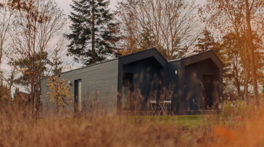 Ein modernes, dunkelgraues Tiny House mit Flachdach und Holzakzenten steht auf einer Wiese, umgeben von Bäumen und hohem Gras im Vordergrund. Die Szene wirkt ruhig und herbstlich. Zwei kleine weiße Stühle und ein Tisch stehen auf einer angedeuteten Terras