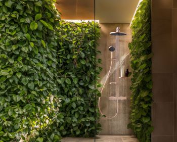 Eine moderne, kupferfarbene Regendusche ist vor einer Wand installiert, die komplett mit grünen Kletterpflanzen bewachsen ist. Das Licht setzt die Duscharmatur und das Grün stimmungsvoll in Szene.