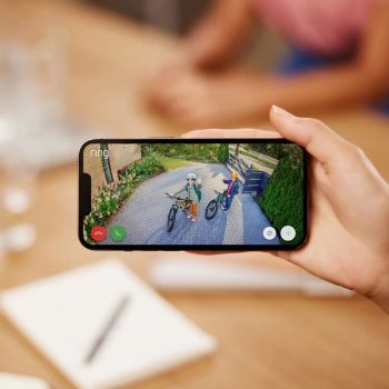 Eine Hand hält ein Smartphone, auf dessen Bildschirm die Live-Ansicht einer gepflasterten Hauseinfahrt zu sehen ist. Auf dem Display sind zwei Personen mit Fahrrädern erkennbar, die in die Kamera winken.