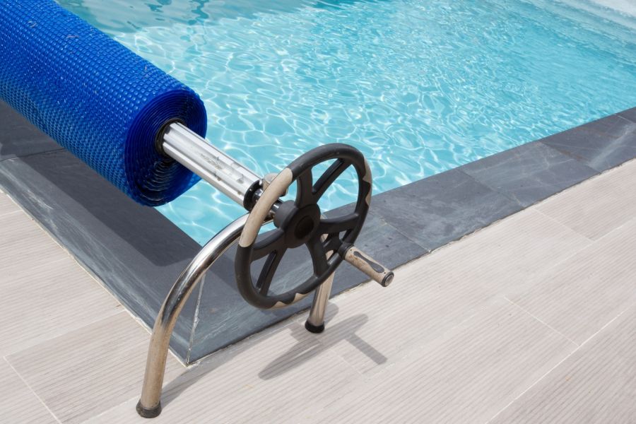 Am Rande eines modernen Swimmingpools mit hellen Fliesen steht eine Aufrollvorrichtung, auf der eine blaue Solarfolie aufgerollt ist. Die Folie dient als Poolabdeckung, um die Wassertemperatur zu halten und Verdunstung zu reduzieren.