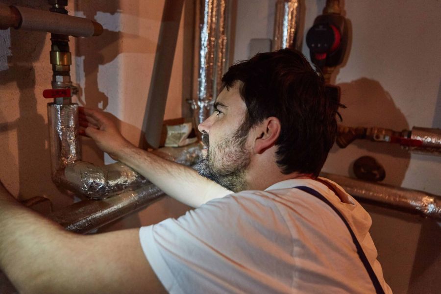 Ein Handwerker in einem Keller isoliert freiliegende Heizungs- und Warmwasserrohre mit einer silberfarbenen Dämmschicht.