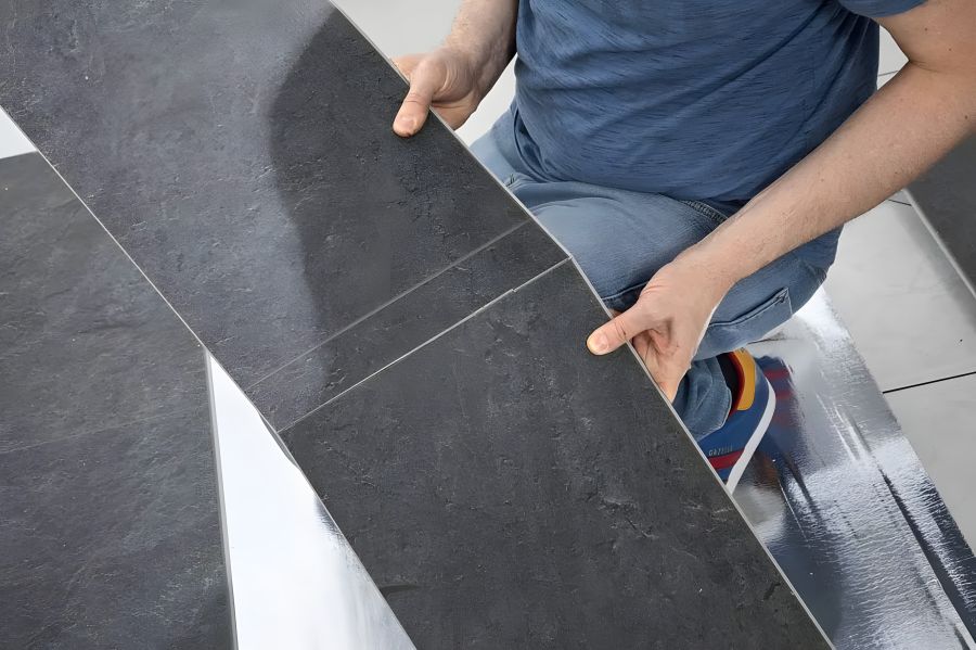 Eine Person in Jeans und blauem T-Shirt verlegt eine Vinylbodenpaneele mit dunkler Steinoptik. Die Hände halten das Paneel in einem Winkel, um es mit dem Klicksystem in das bereits verlegte Element einzufügen. Im Hintergrund liegt eine silberne Trittschal