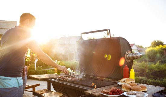 Ein Mann steht an einem großen Barbecue-Grill im Freien, während die warme Abendsonne im Hintergrund scheint. Er wendet Fleisch auf dem Rost mit einer Zange und hält in der anderen Hand eine Bierflasche. Auf einem Holztisch daneben stehen Burgerbrötchen,