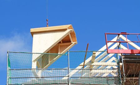 Eine große, vorgefertigte hölzerne Dachgaube hängt an einem Kranhaken über einer Baustelle mit grünem Sicherheitsnetz vor blauem Himmel.
