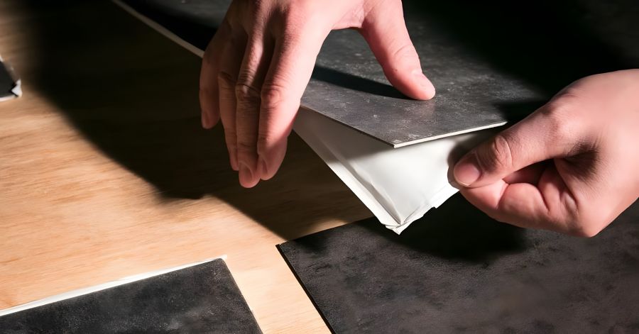 Eine Nahaufnahme zeigt zwei Hände, die eine selbstklebende Vinylbodenfliese mit dunkler Steinoptik von der Schutzfolie ablösen. Der Boden wird auf eine Holzunterlage vorbereitet.
