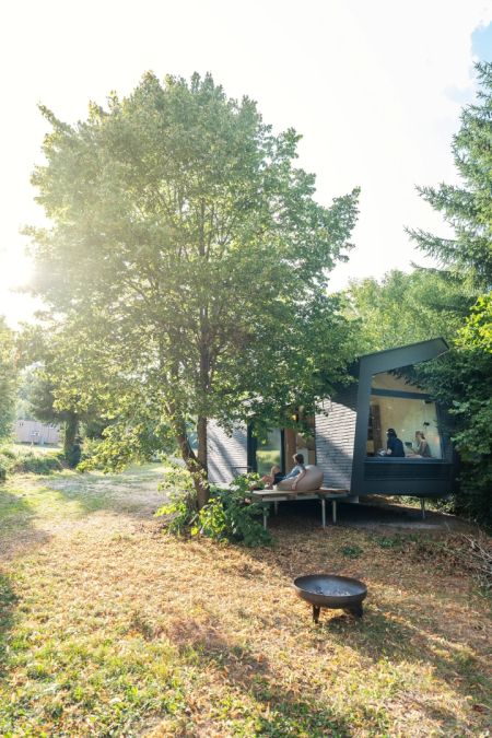 Ein modernes, winkliges Tiny House steht unter einem großen Baum. Auf der Holzterrasse sitzen Menschen und entspannen. Im Vordergrund ist eine Feuerschale auf einer Wiese zu sehen. Das Haus hat eine große Glasfront.